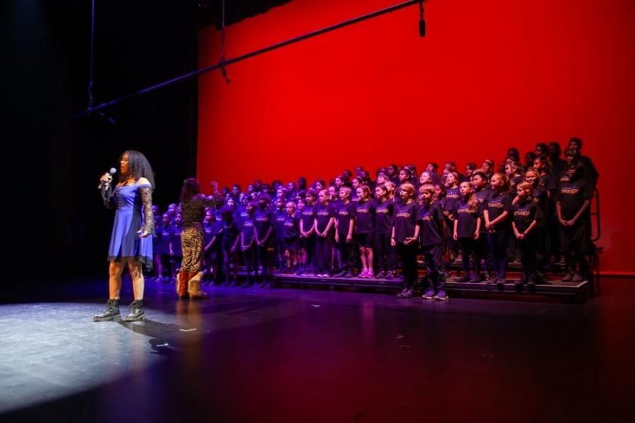 A woman is singing into a microphone on a stage in front of a large choir.