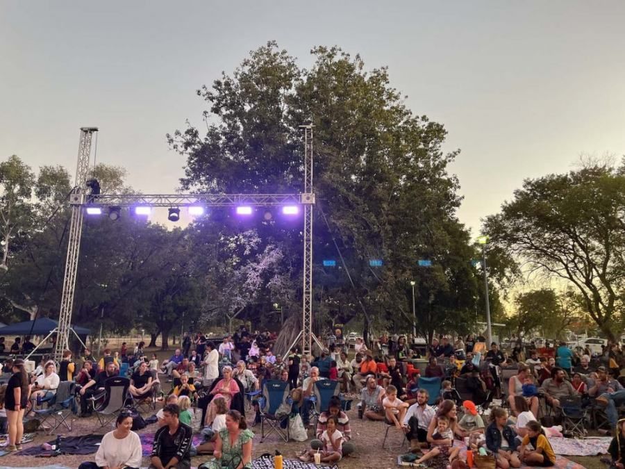 A large group of people are sitting in front of a stage in a park.