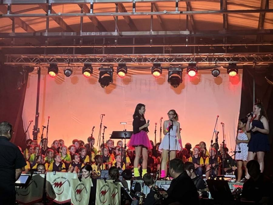 A group of girls singing on a stage in front of an orchestra