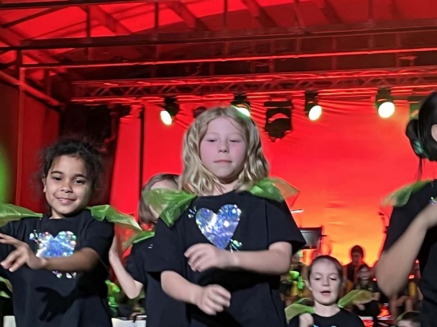 A group of young girls wearing black shirts with a heart on them are dancing on a stage