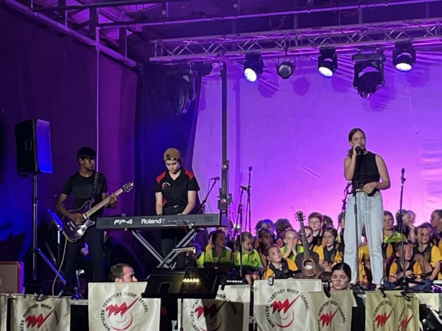 A woman is singing into a microphone on a stage with a band behind her.