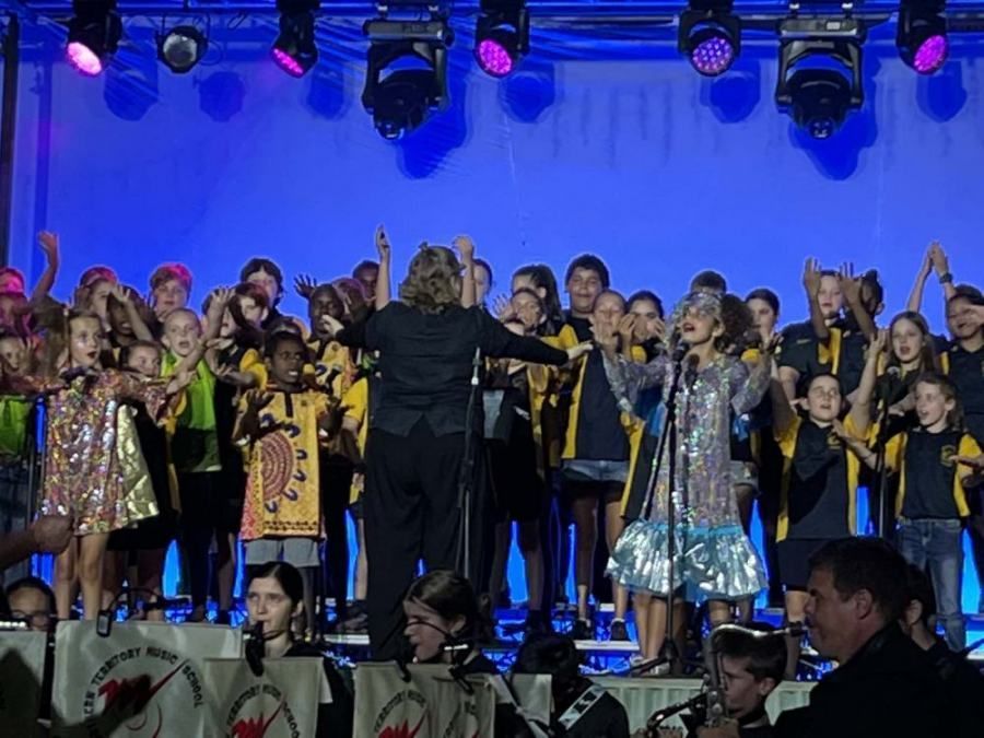 A group of children singing on a stage with a conductor