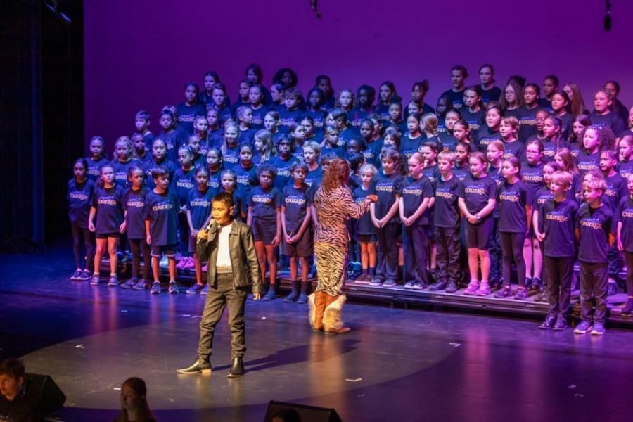 A man is singing into a microphone in front of a large choir on a stage.