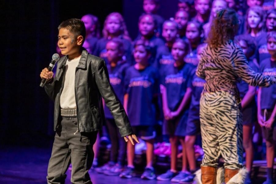 A boy is standing on a stage holding a microphone in front of a choir.