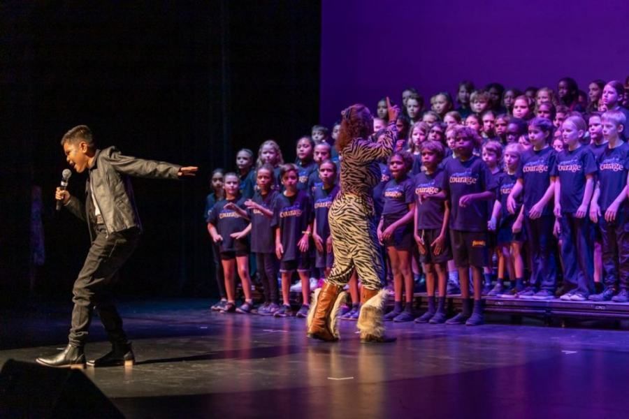 A man is standing on a stage with a microphone in front of a choir of children.
