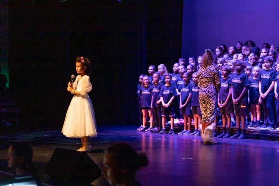 A little girl is standing on a stage with a microphone in front of a choir.