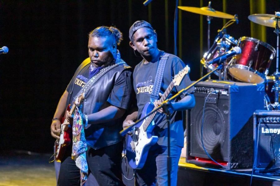 Two men are playing guitars on a stage in front of a drum set.