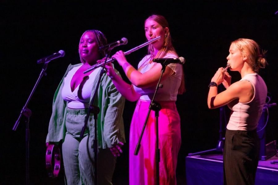 Three women are playing instruments on a stage and singing into microphones.