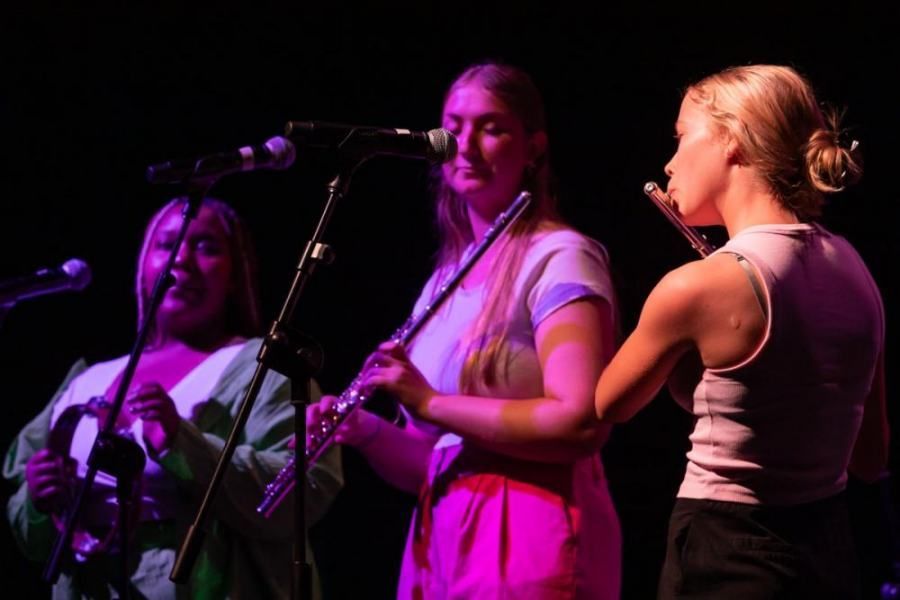 Three women are playing instruments and singing into microphones on a stage.