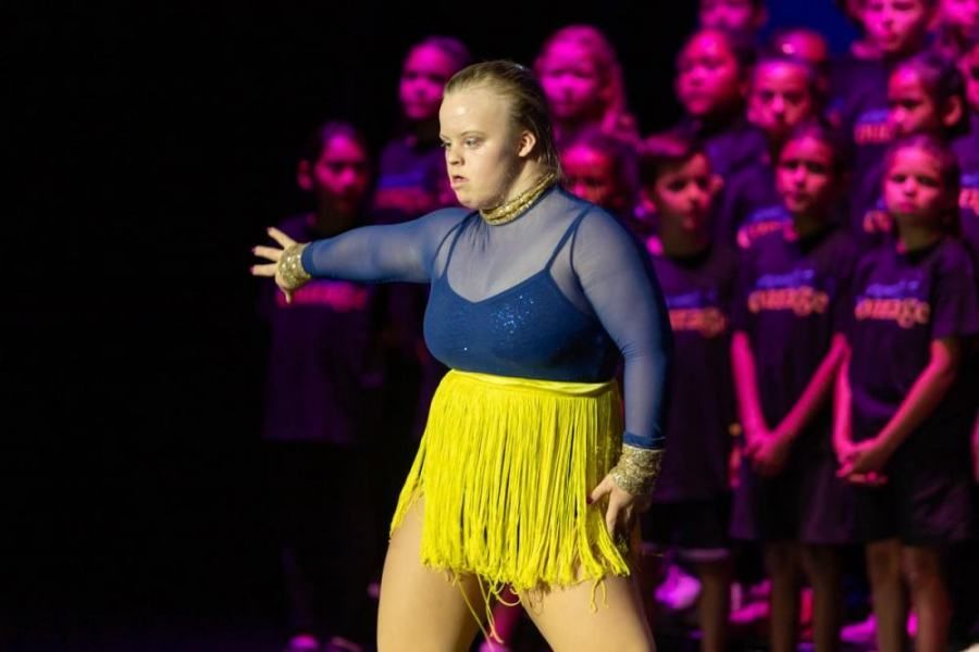 A woman in a blue top and yellow skirt is dancing on a stage in front of a choir.