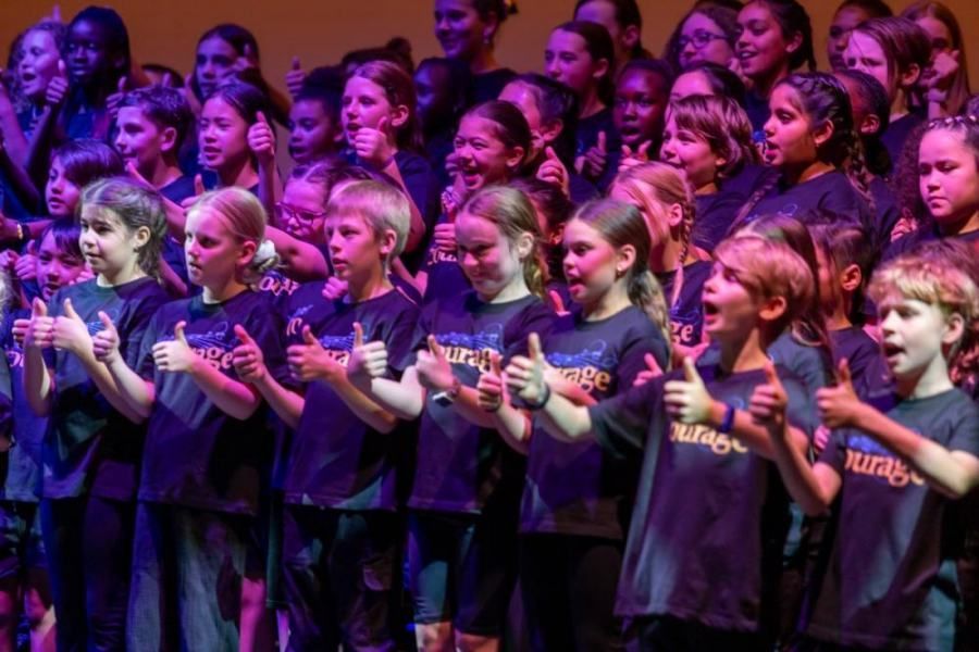 A large group of children are singing and giving a thumbs up.