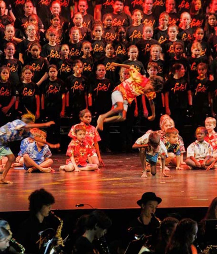 A group of children are performing on a stage in front of a large choir wearing shirts that say 