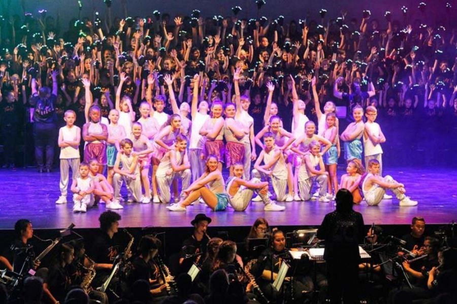 A large group of children are dancing on a stage in front of an orchestra.