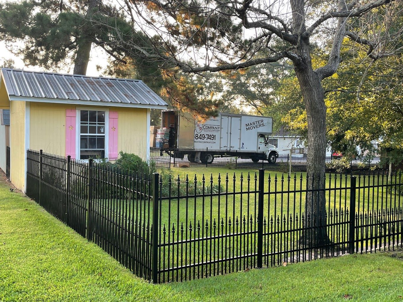 A white truck is parked in front of a yellow shed behind a black fence.