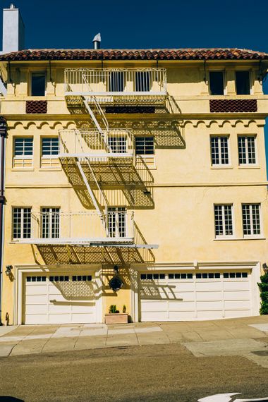 Yellow stucco apartment building with white fire escape, two garage doors, and blue sky.
