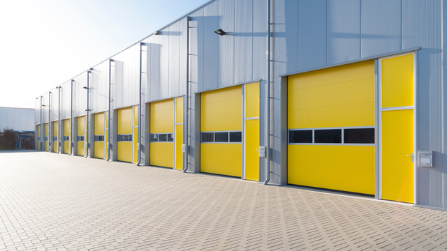 Row of yellow industrial doors on a warehouse with a gray exterior, on a brick paved surface.