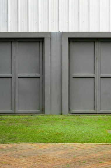 Two grey doors set in a grey wall, in front of grass, with a corrugated white metal background.