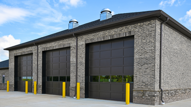 Brick building with three garage doors. Brown doors, yellow bollards, blue sky.