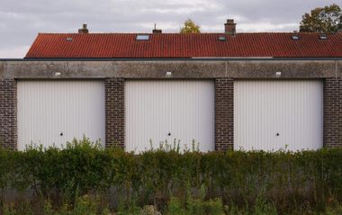 Three white garage doors in a row, with a red-tiled roof above, fronted by a hedge.