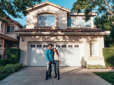Couple standing in front of a two-story beige house with a two-car garage. Blue sky.