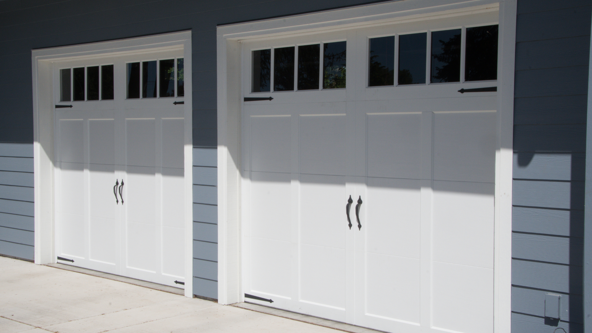 Two white garage doors with black handles and window panels, set in a blue siding house.