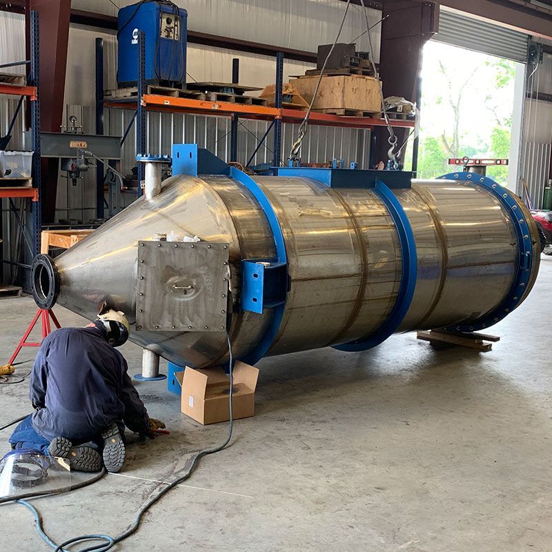Worker welding a large metal cylindrical tank in a workshop, with blue support rings and equipment nearby