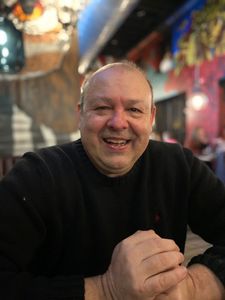 A person with a smiling expression, wearing a black sweater, sits with hands clasped in an indoor setting.
