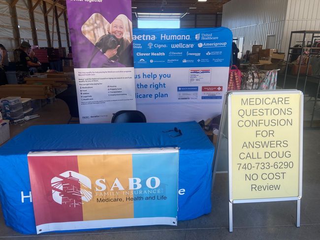 A Sabo Medicare information booth with a blue table, branded banners, and a sign with contact details for inquiries.