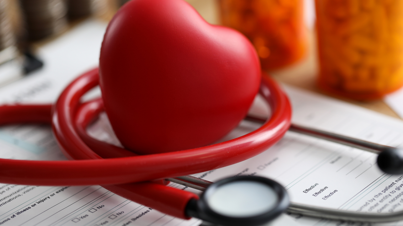 A red model heart rests on a red stethoscope, placed atop medical paperwork near orange pill bottles.