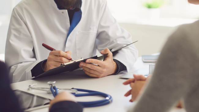 A doctor writes on a clipboard during a consultation with a patient in a medical office, with a stethoscope on the table.
