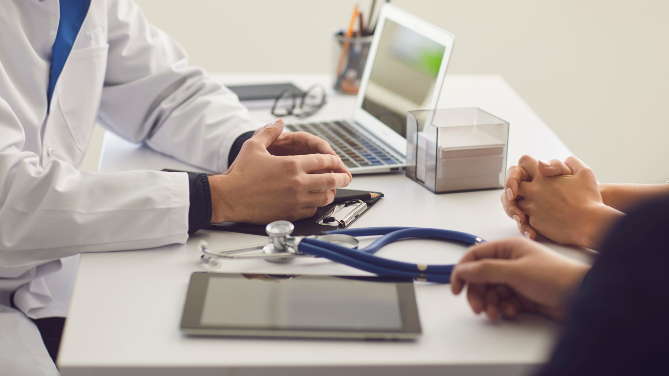 A doctor sits at a desk with a laptop and stethoscope, consulting with a patient in an office setting.
