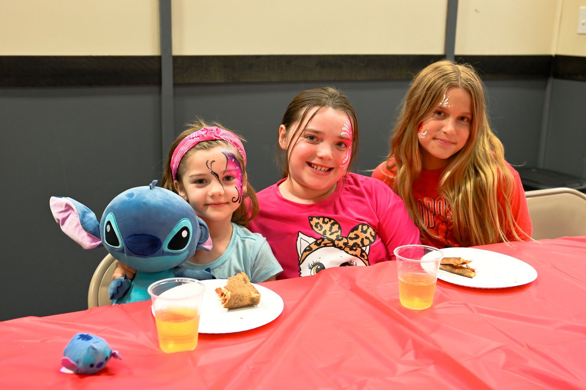Three young girls are sitting at a table with plates of food and a stuffed animal at Impact Fun Zone