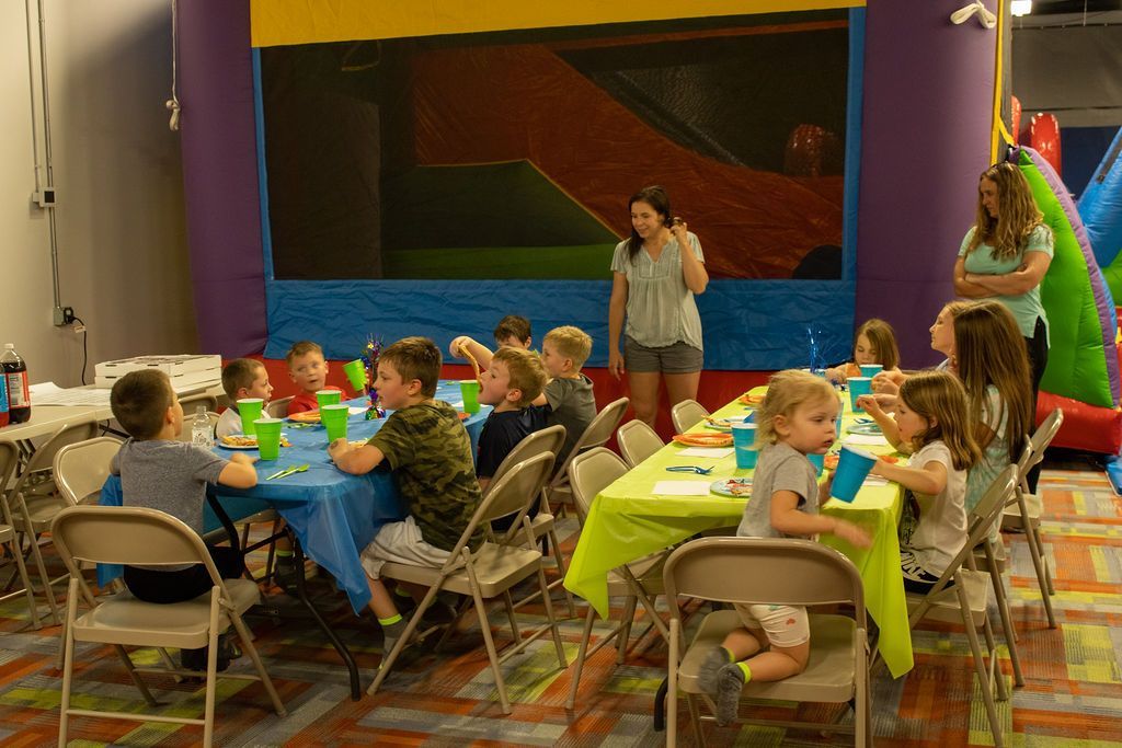 A group of children are sitting at tables at a birthday party in front of a bouncy house at Impact Fun Zone