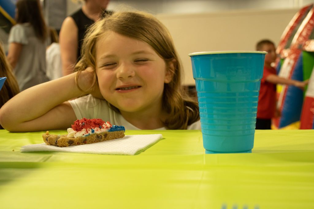 A little girl is sitting at a table with a piece of cake and a cup  Impact Fun Zone