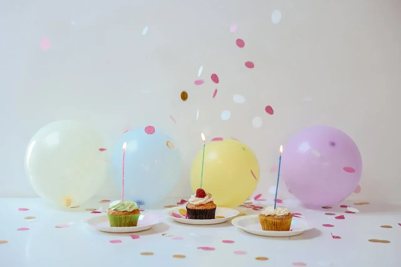 Three cupcakes with candles on them are sitting on plates on a table with balloons and confetti.