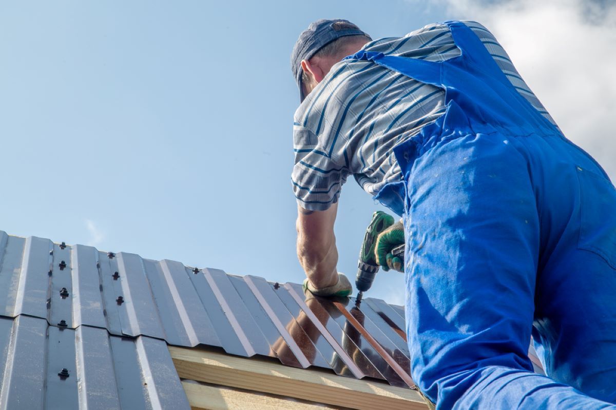 A Man in Blue Overalls is Working on a Metal Roof — Hume Plumbing & Roofing Wollongong in Dapto, NSW