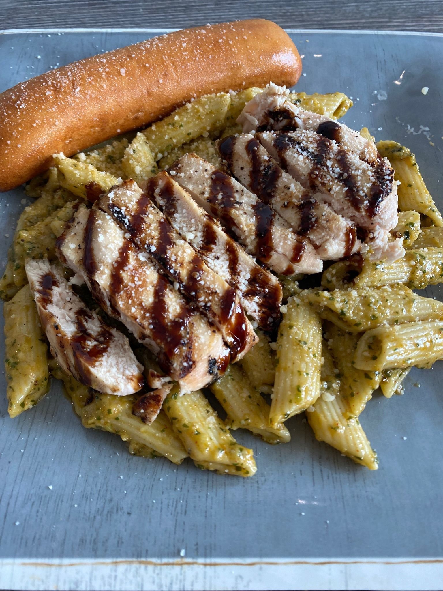 Grilled chicken over penne pasta with green sauce, topped with balsamic drizzle and a breadstick on a gray plate