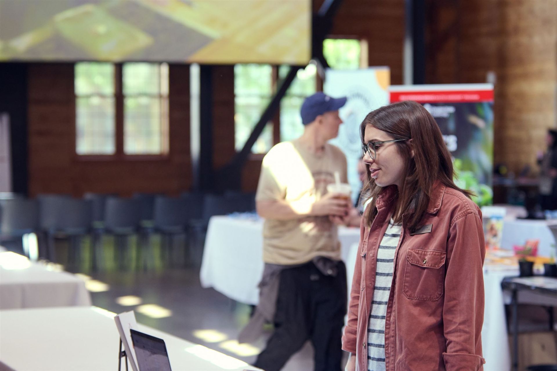 Woman in glasses smiles, stands near tables in a hall with a man in the background.