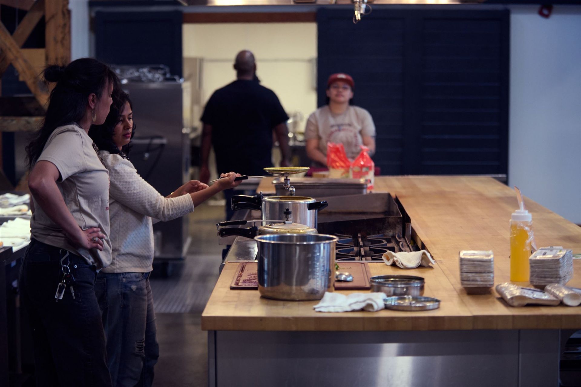 Two women in a kitchen, pointing at cooking area. A chef works in the background.