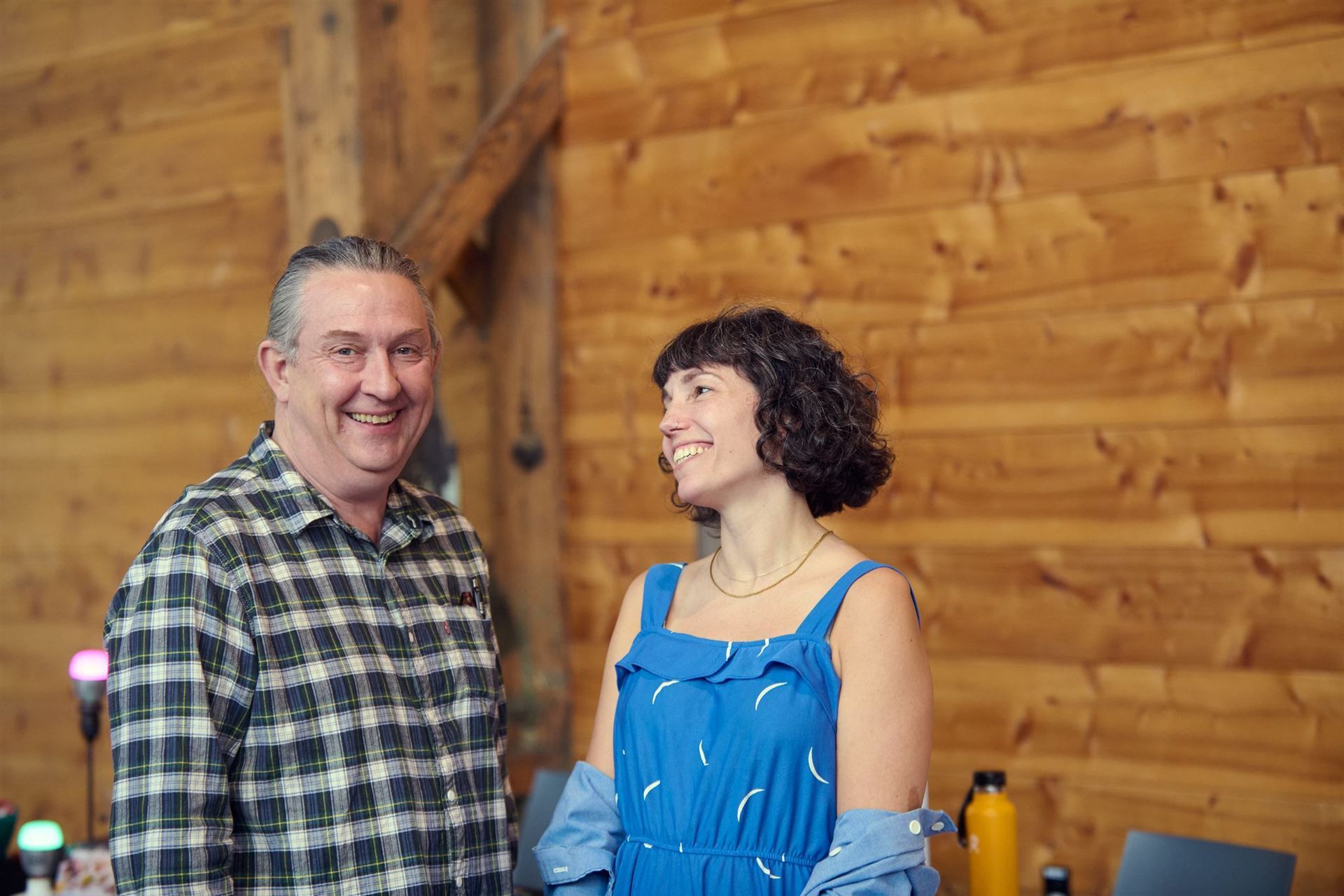 Man in plaid shirt and woman in blue dress smile in front of wood wall.