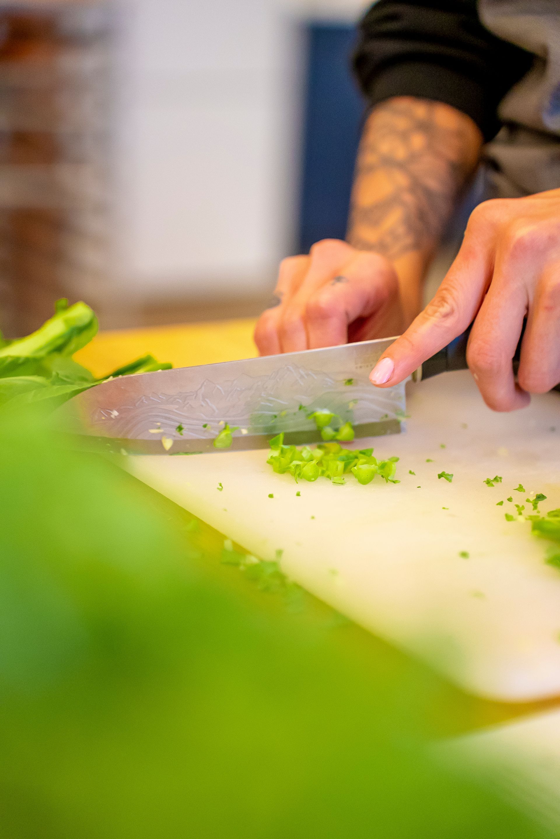 Person chopping green vegetables on a white cutting board, showcasing tattooed arm and hands.
