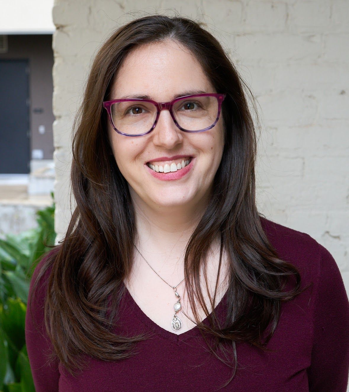 Woman with glasses smiles, wearing a burgundy sweater, standing near a white brick wall.