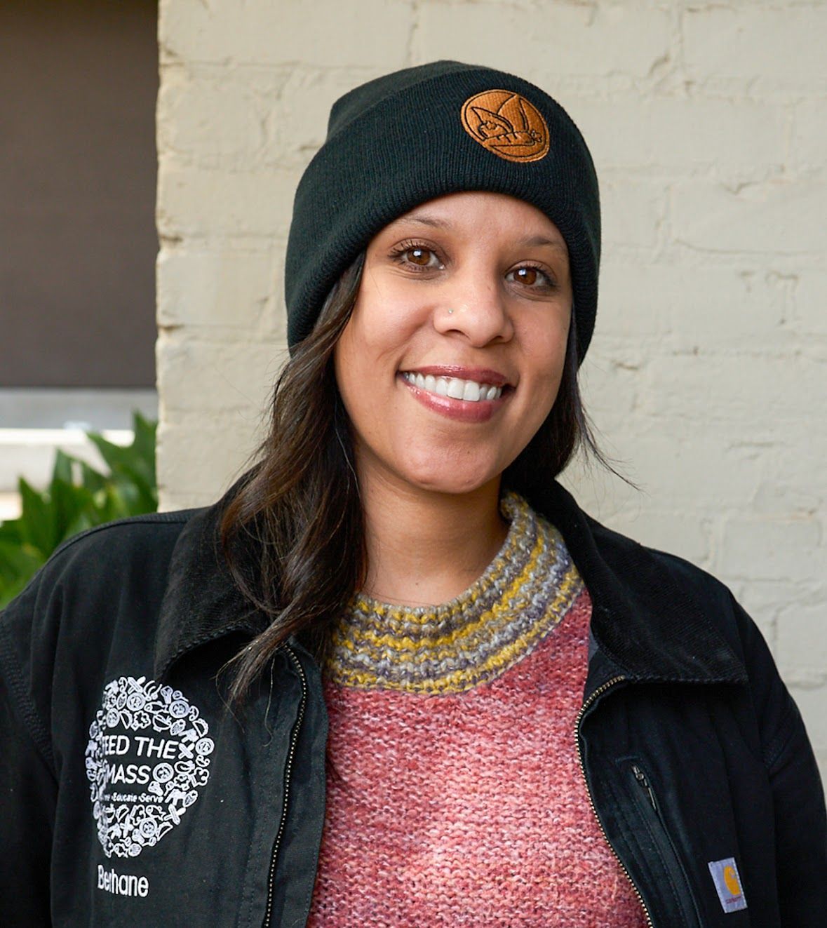 Woman in black hat and jacket smiles, standing outside.