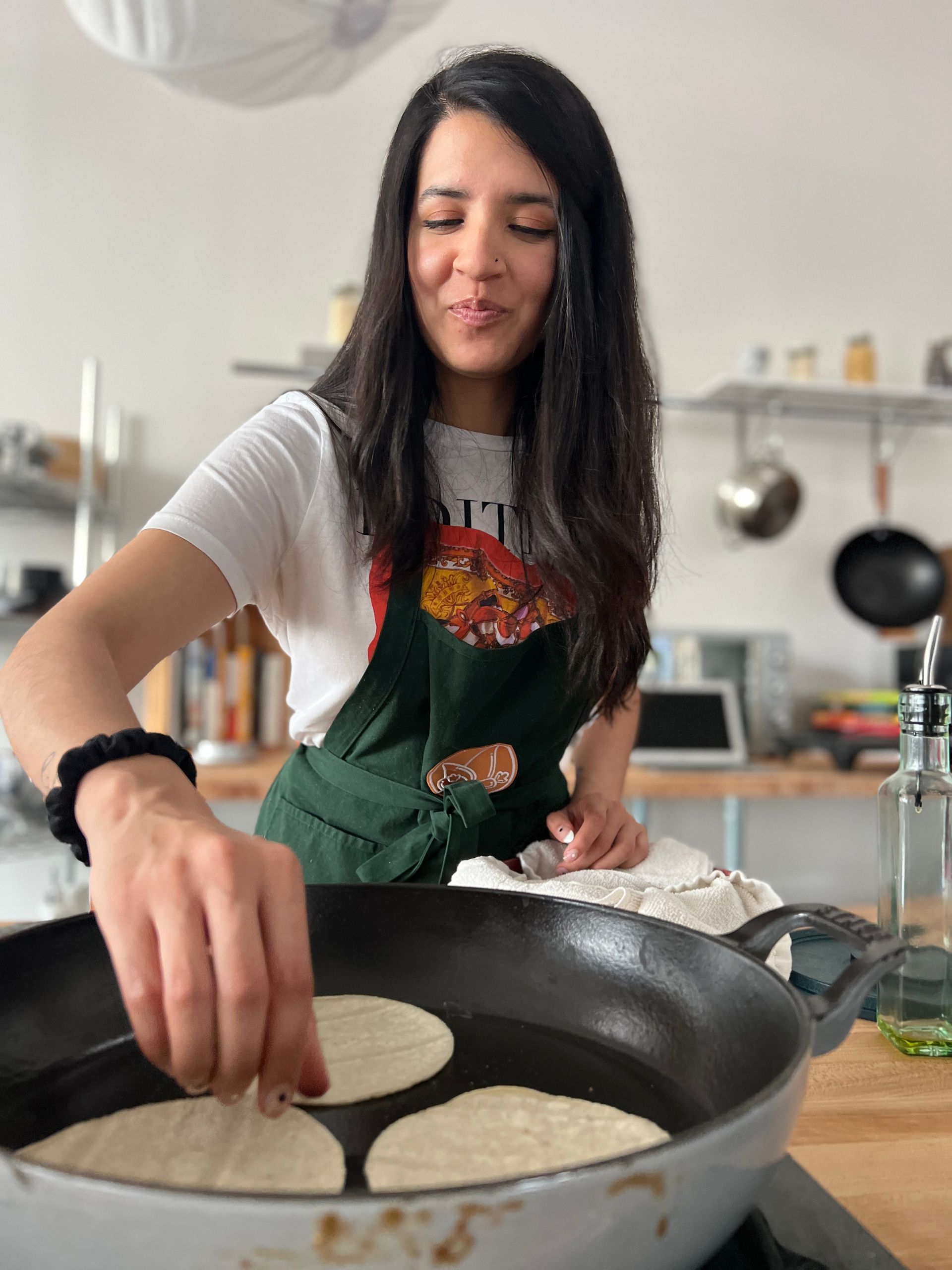 Woman in apron frying tortillas in a pan, smiling in a kitchen.