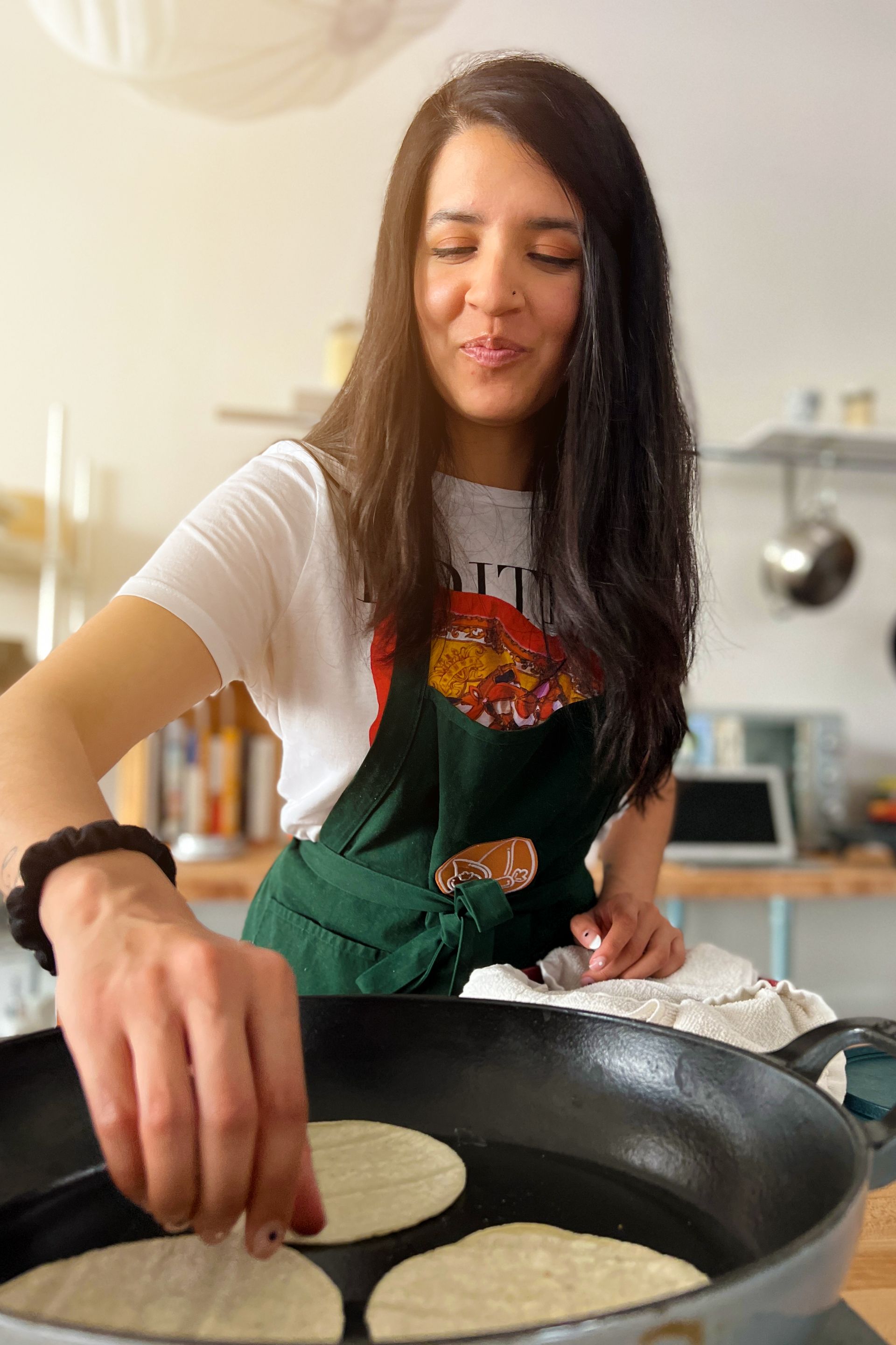 Woman in apron frying tortillas in a pan. Bright kitchen setting; she's smiling and reaching in.
