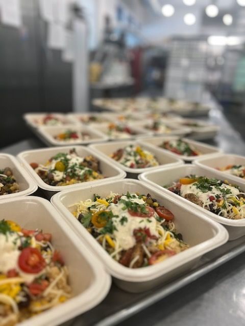 Prepared meals in white containers on a metal surface, likely in a commercial kitchen.