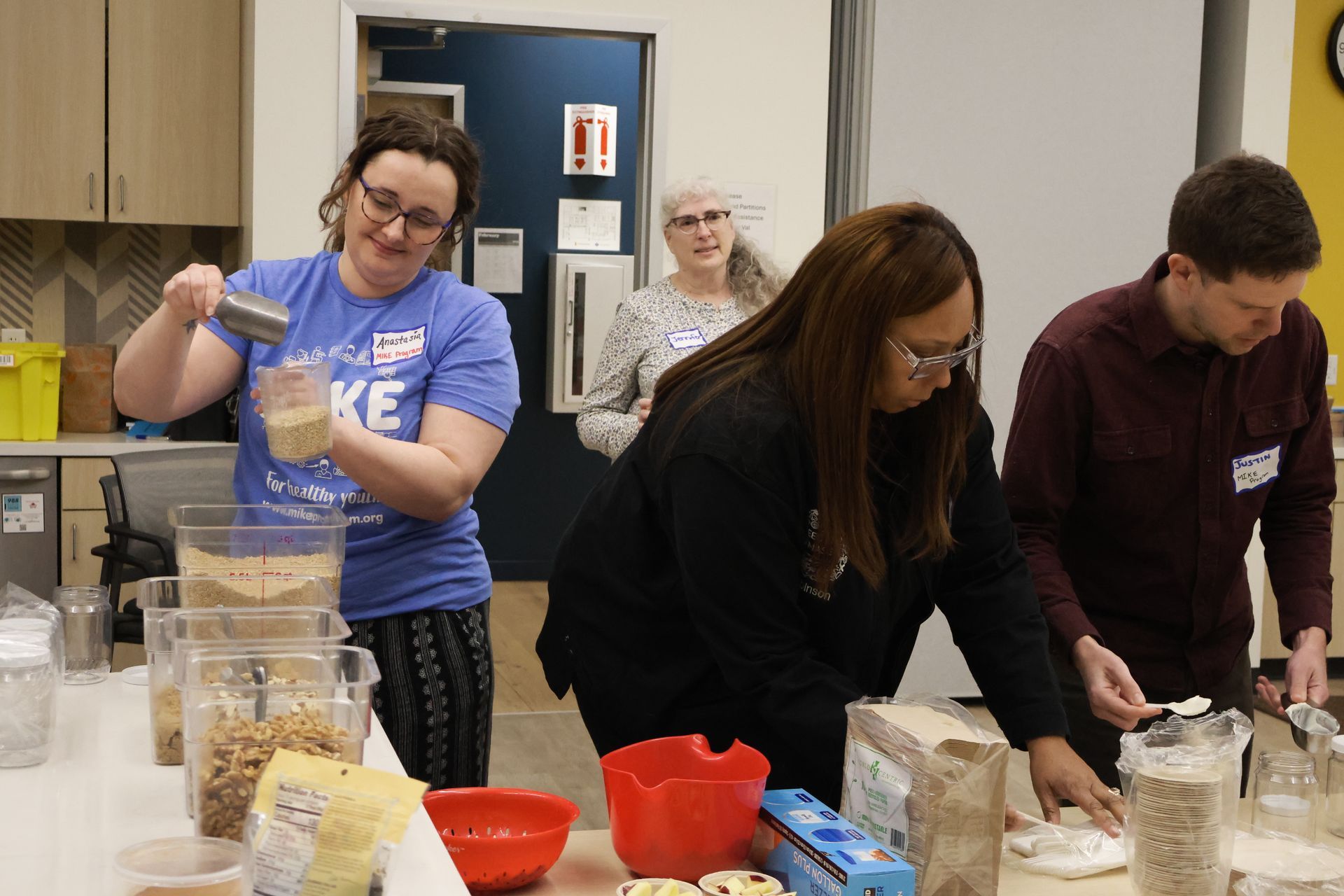 People preparing food in a kitchen. One woman pours ingredients, others measure and work.