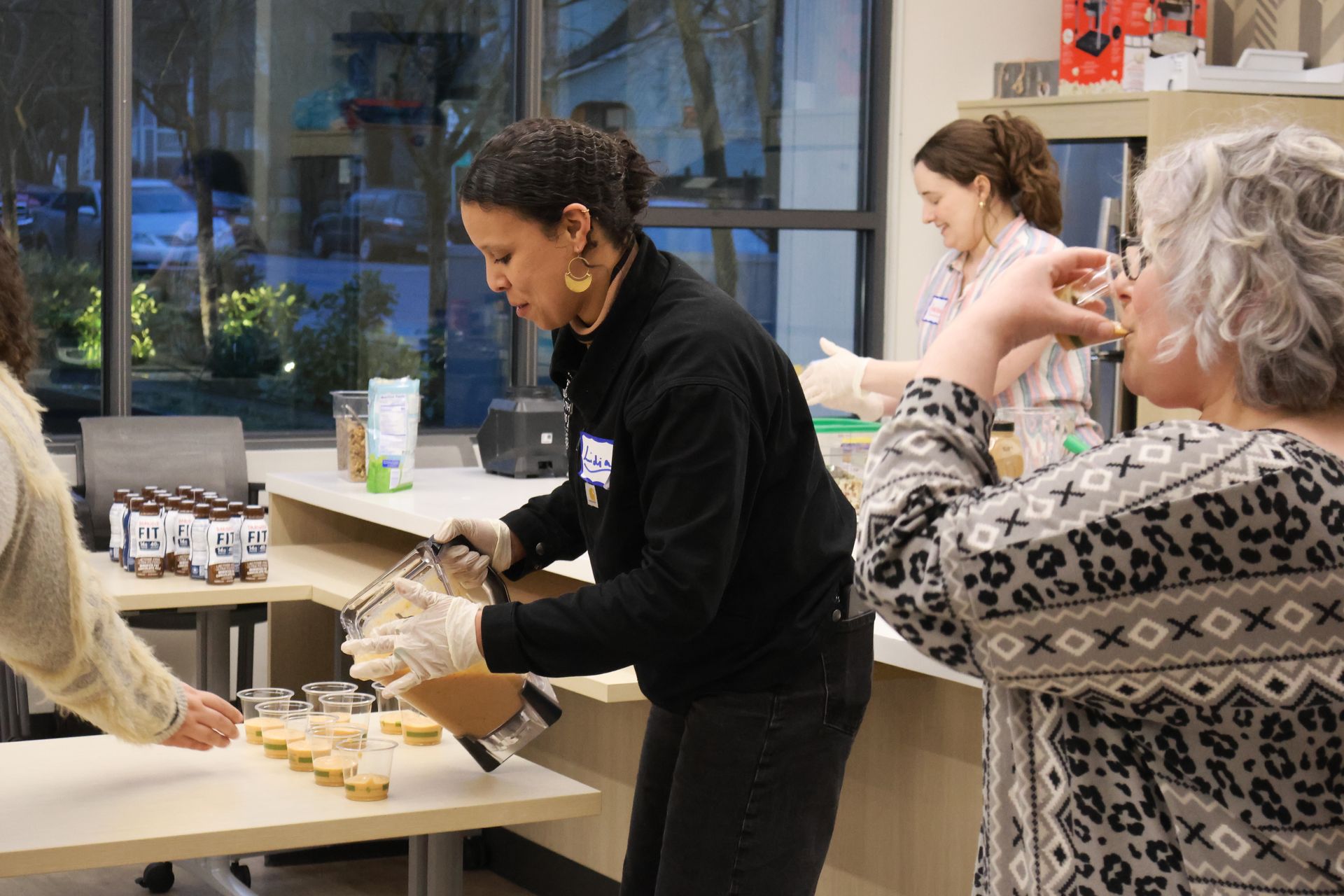People preparing food at a table, one pouring a liquid from a bowl. Indoor setting.