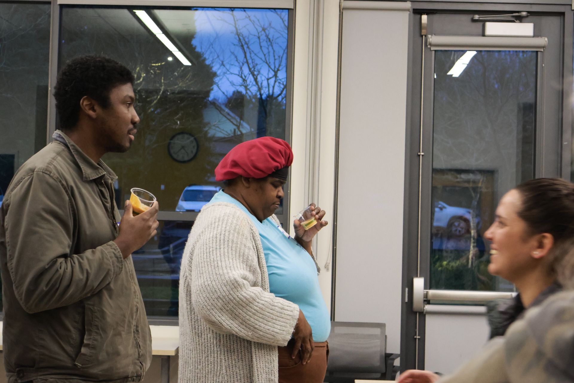 Three people in a room with a large window.  Two people hold drinks, one wears a red head covering. A woman smiles.
