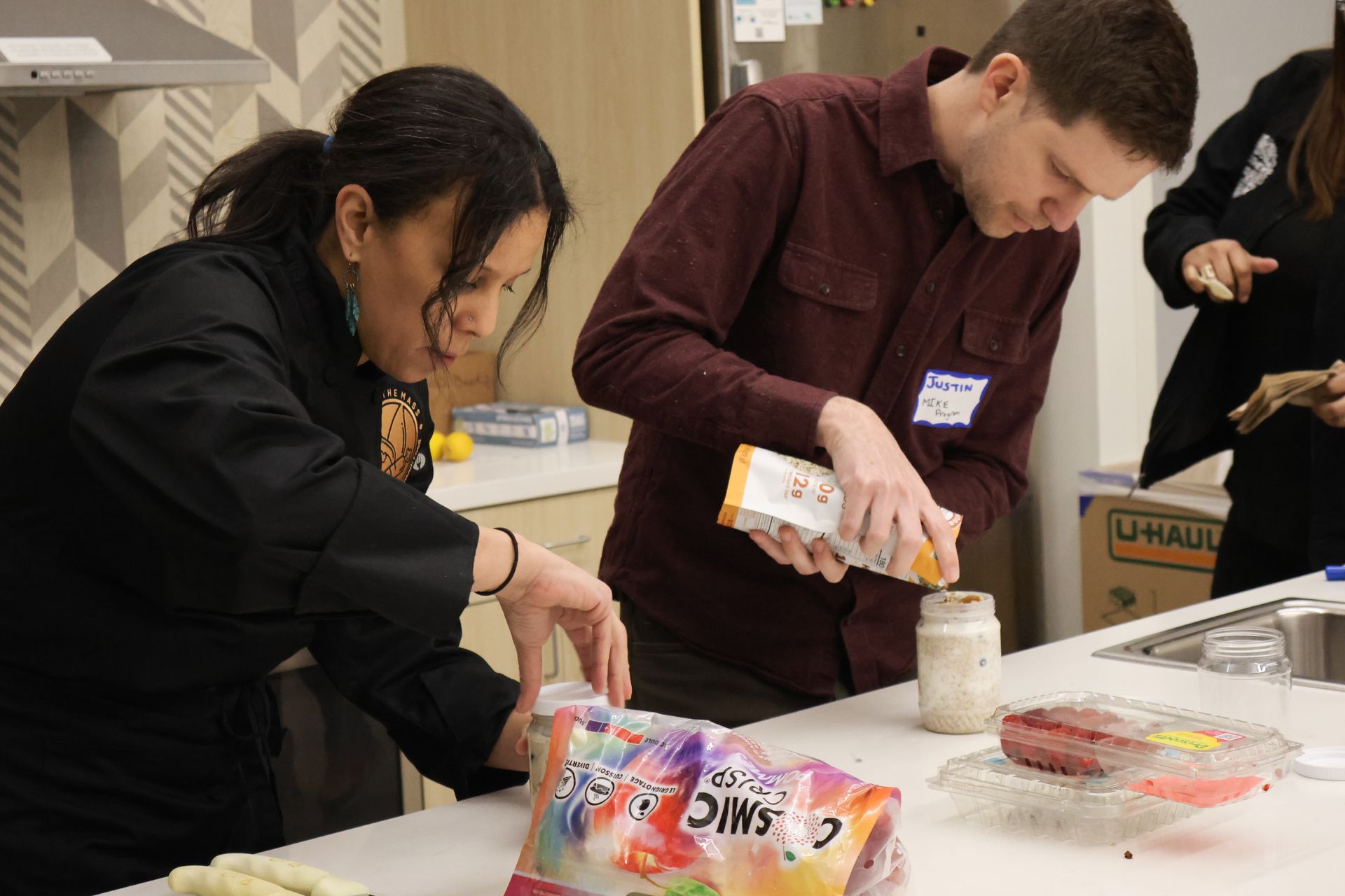 Two people in a kitchen preparing food, one pouring from a bag, the other working with a plastic bag.